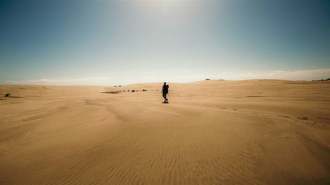 A person with a backpack walks across sand dunes under a bright sun.
