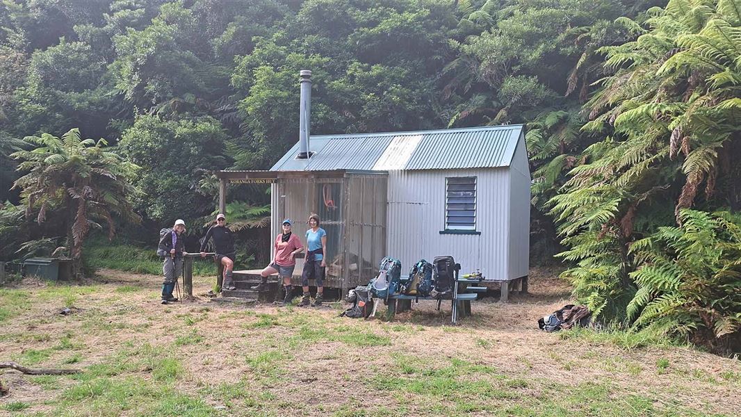 Four people standing outside a hut with packs laying on a picnic table. 