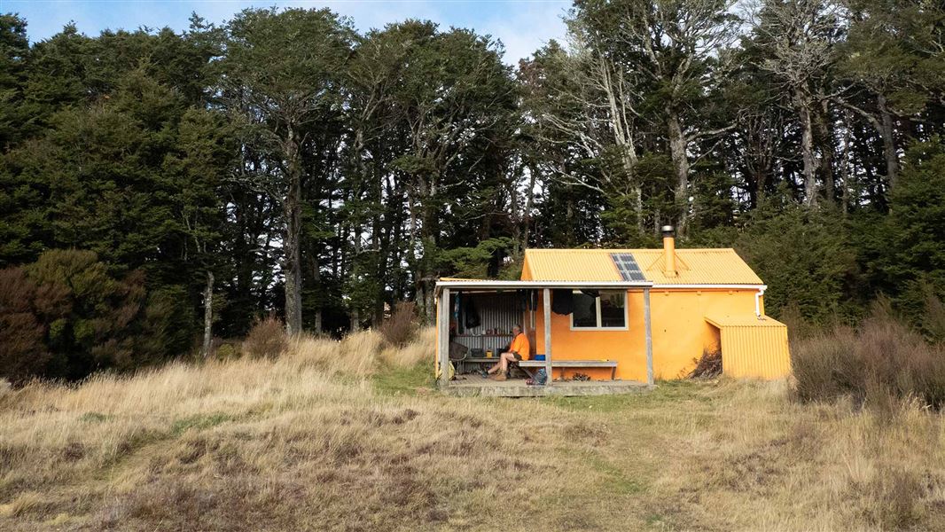 An orange hut sitting on a grassy rise with tall trees behind it. A man in hi vis sits on the deck. 