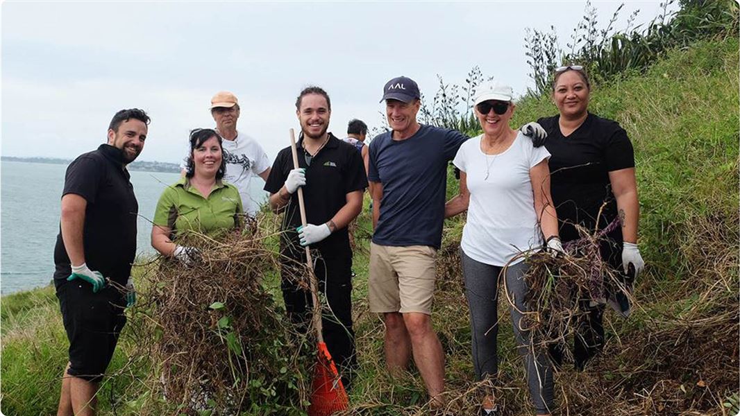 A groupf of seven adults on a grassy bank taking a break from picking up weeds to pose for the camera. 
