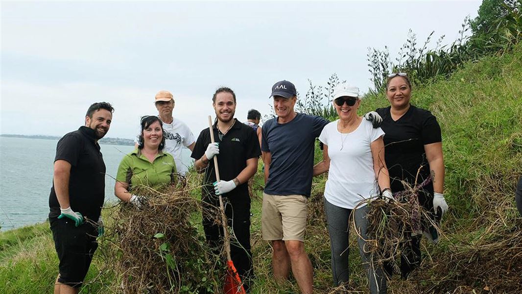 Group of volunteers with a DOC ranger weeding on a hill siden with the ocean in the background.
