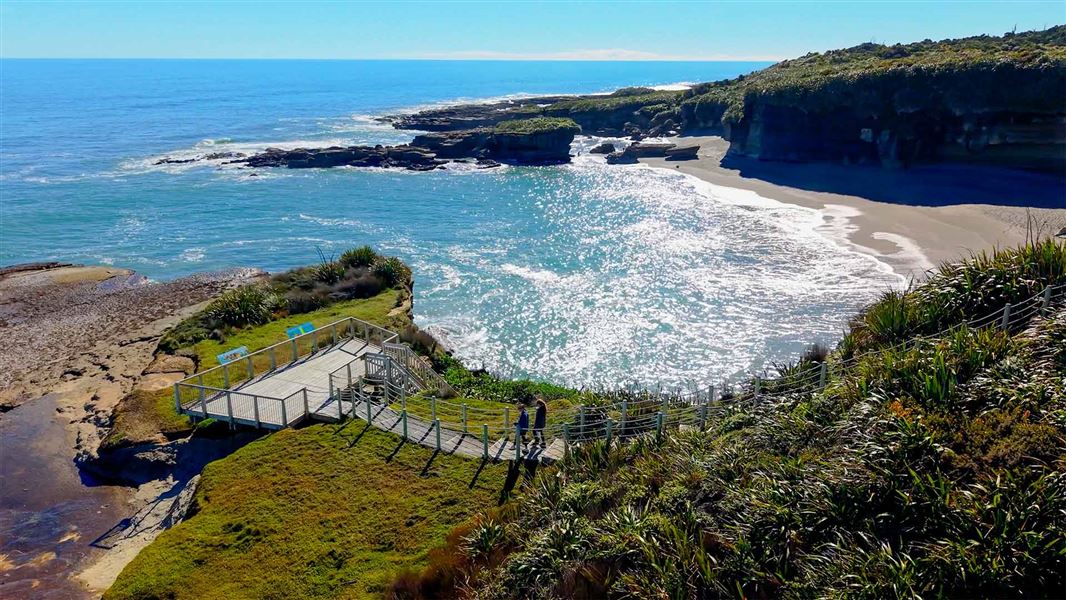 Aerial view of two walkers descending steps to a wooden platform above sparkling sea and a cove ringed by cliffs.