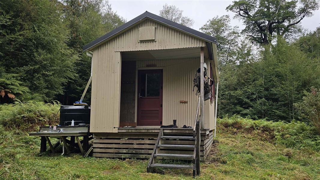 A small corrugated beige hut with a small black water tank beside it and a small porch with gumboots on it. 