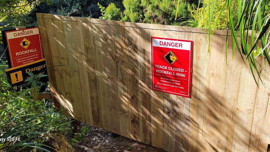 A wood wall, festooned with warning signs, blocks a path.