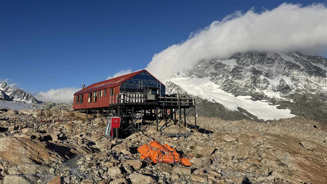 A deep red hut atop rocks with snowy peaks behind covered by a long cloud and big blue sky beyond. 