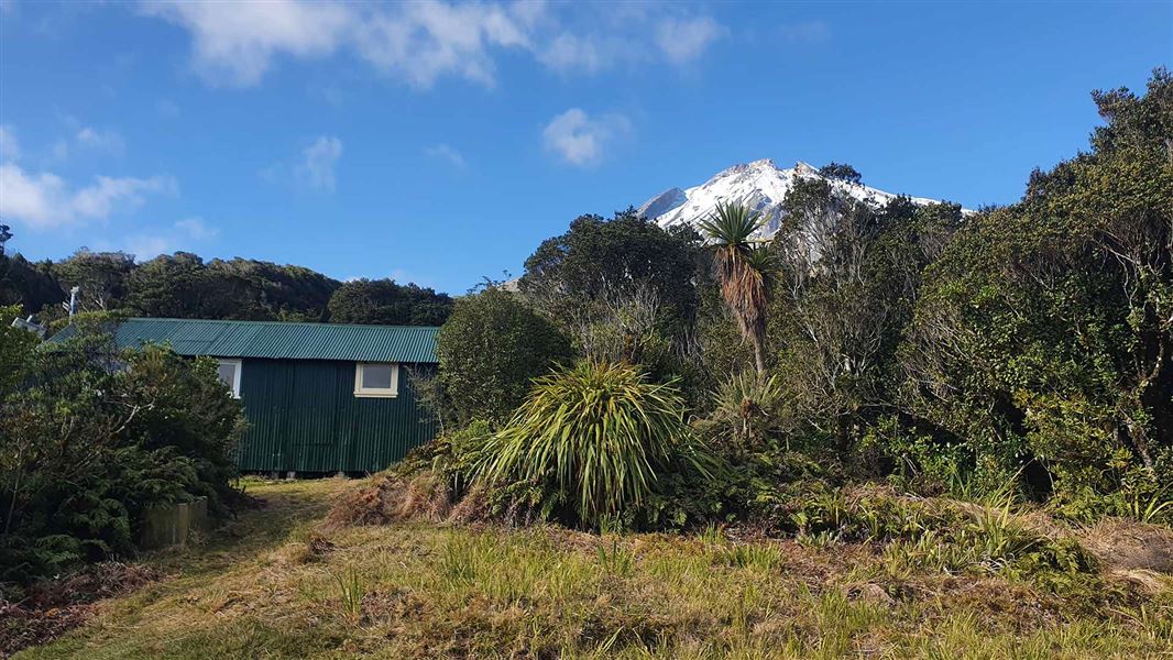 A brown hut nestled in front of bush and a snow capped peak to its right at the rear. 