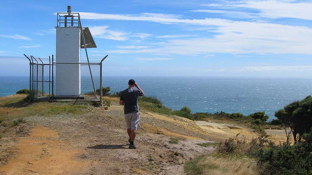 A person walks toward a small rectangular lighthouse on the top of a seaside ridge.
