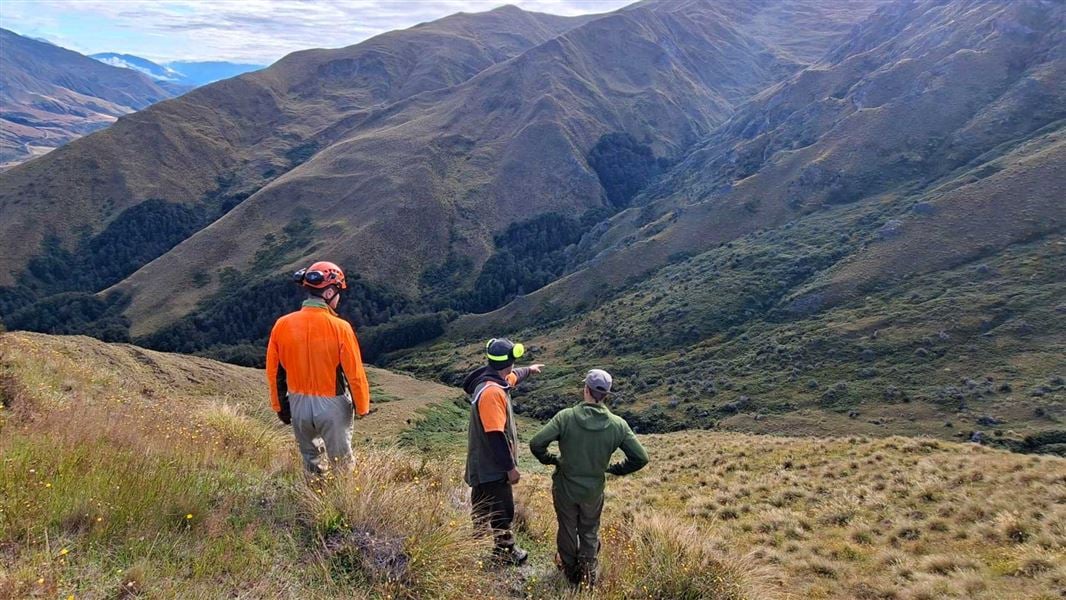 Three staff members standing on a hill wearing bright orange and green and observing the hills beyond them. 
