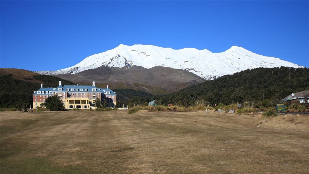 A wide shot of flat lawn with the chateau nestled in front of hills with a snowcapped mountain behind them. 