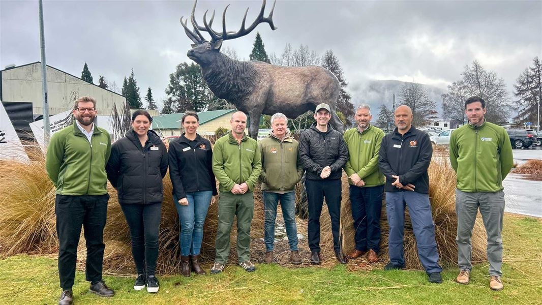 Nine people stand outdoors in front of a large bronze elk (wapiti) statue on a cloudy day. The group is lined up on a grassy area with tall ornamental grasses in the foreground, wearing outdoor jackets and casual clothing. 
