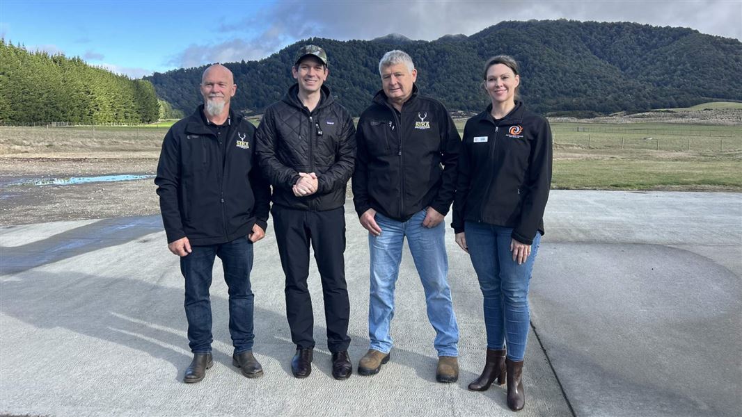 Four people stand on a concrete pad in a rural landscape, with grassy paddocks, pine trees and forest-covered hills in the background under a cloudy sky.