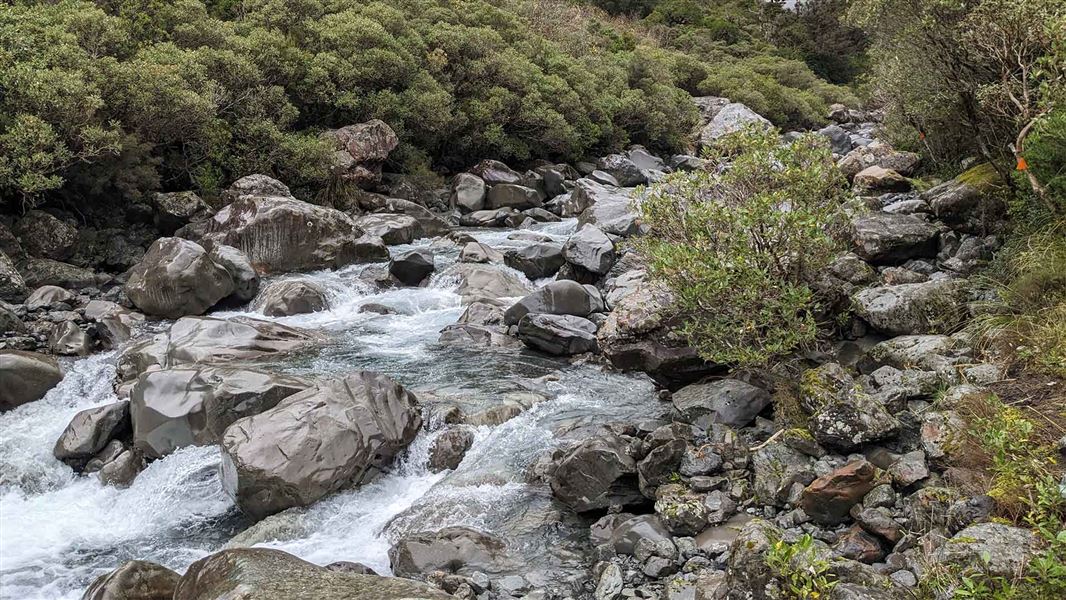 A swift river flowing through a rocky valley. Orange triangles mark a route along the edge of the river.