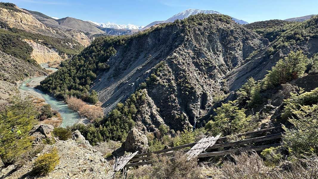 River winding through a steep rocky valley with forested slopes and snow-capped mountains in the distance. 