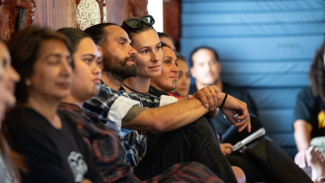 A group of people sit against the wall of a marae during a course on the Māori language and the environment.