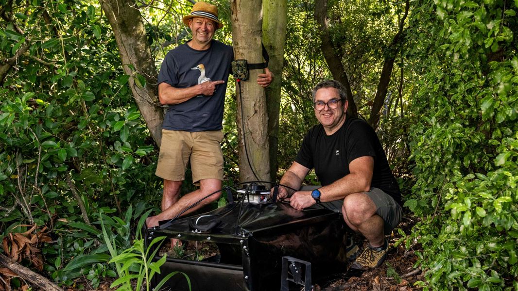 Two men stand in the forest next to an automated trapping system.
