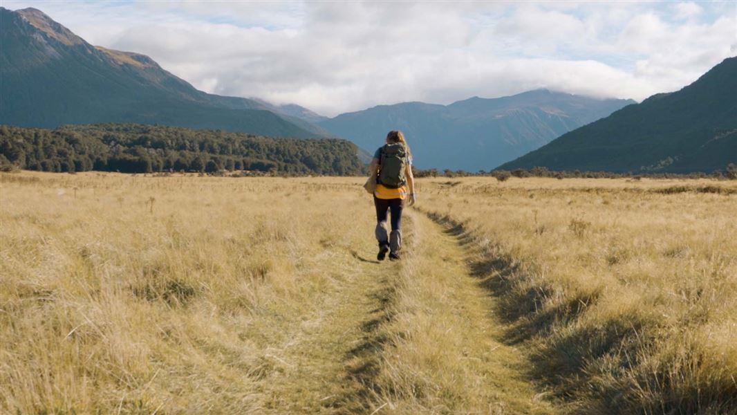 A person walks down a grassy path in a valley surrounded by mountains.