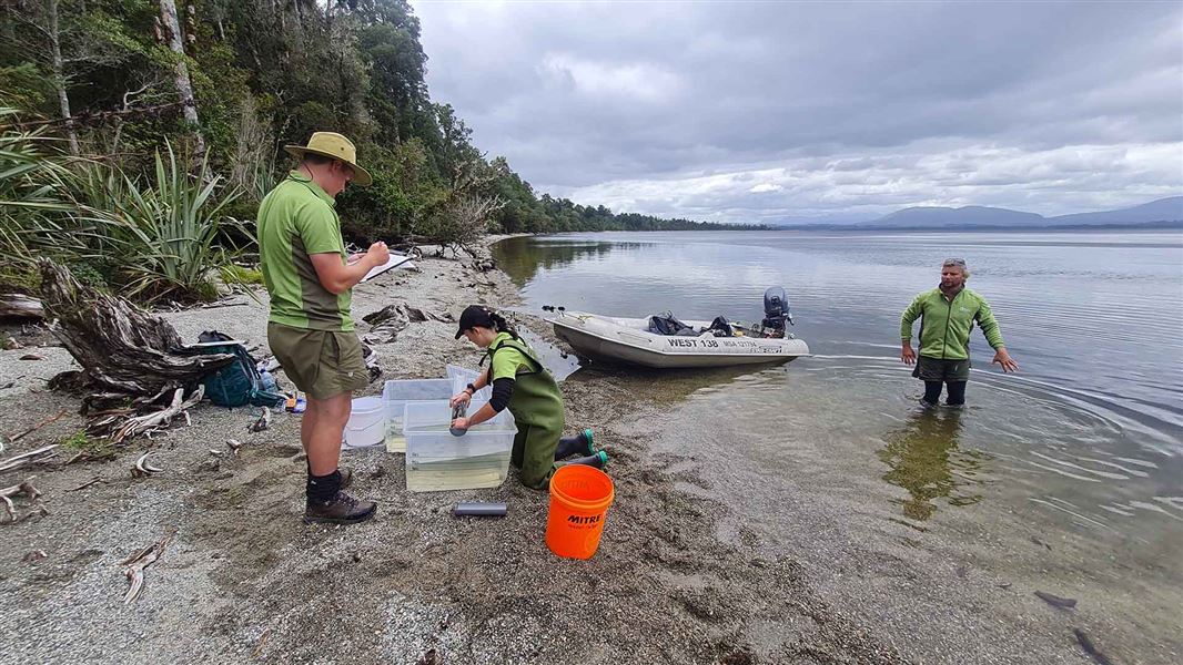 Three people working on the shore of a lake. Equipment, including a small boat, are in view.
