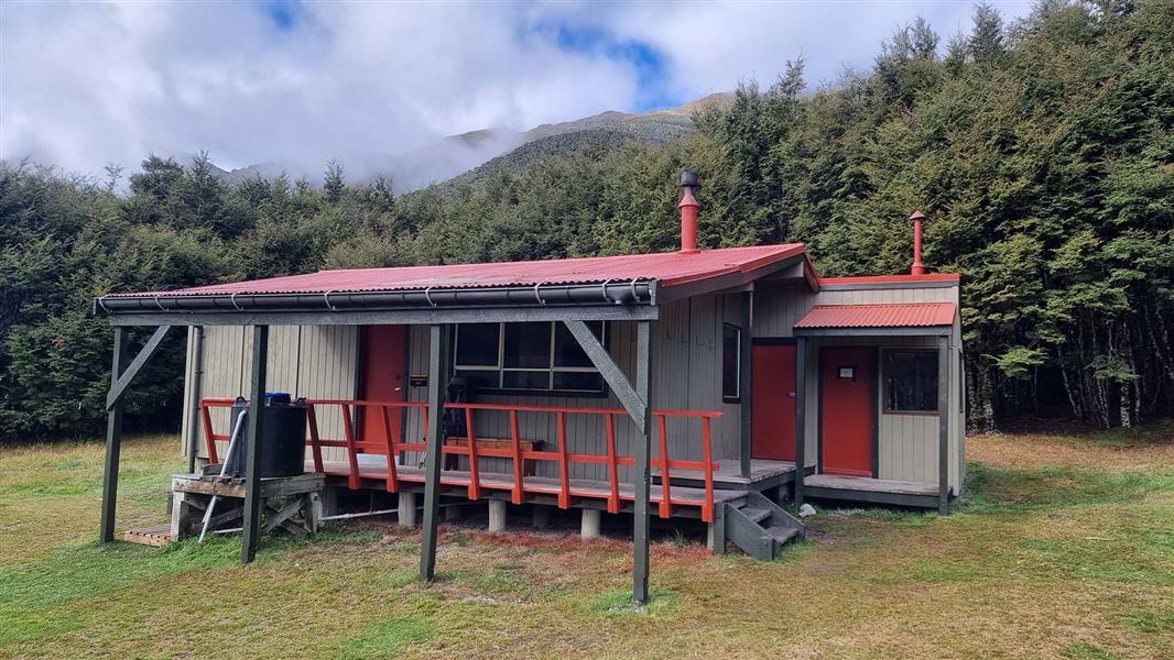 A well-maintained looking hut with thickly wooded hills in the background.