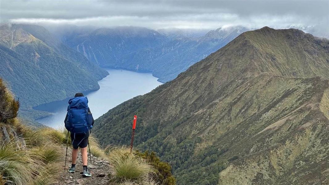 Tramper on a narrow path looks down at a lake ringed with step hills.