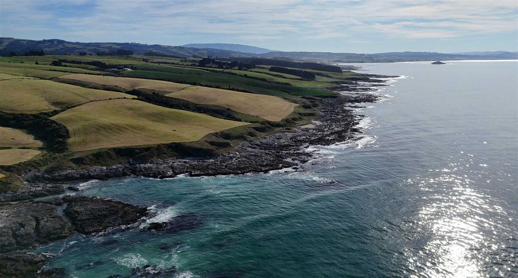 Wide coastal landscape showing rolling farmland meeting a rocky shoreline and calm ocean under a partly cloudy sky.