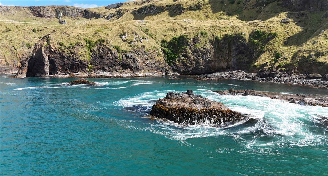 Rocky coastal cliffs with dry grass above, turquoise water below, and waves breaking around scattered offshore rocks.